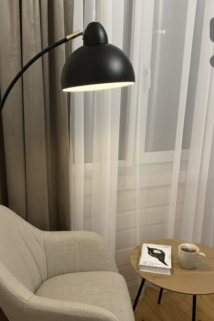 Крупний план зони відпочинку. Світле текстильне крісло стоїть біля круглого дерев’яного столика, на якому лежить розгорнута книга та чашка. Поруч — високий чорний торшер. На фоні — легка біла тюль та щільні штори кольору капучино.